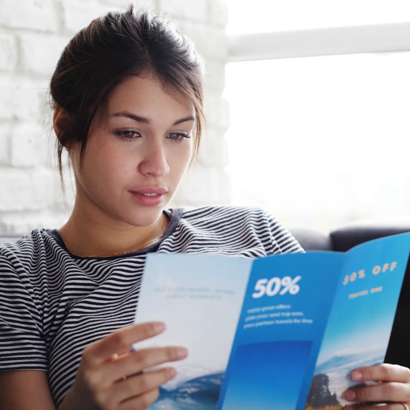 A woman in a striped t-shirt sitting on a sofa reading a blue 50% off advertising leaflet.