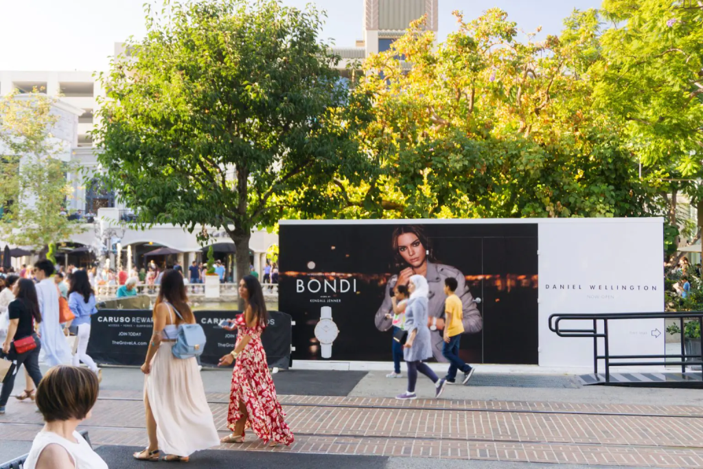 People walk past a large outdoor advertising display featuring a model and various products. Trees and a building are in the background.