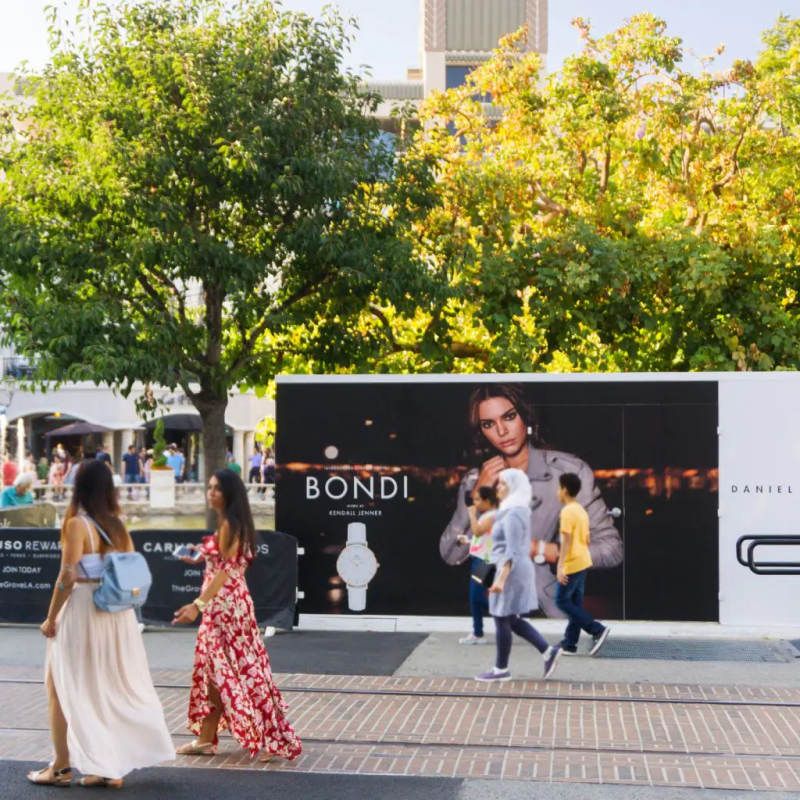 People walk past a large outdoor advertising display featuring a model and various products. Trees and a building are in the background.