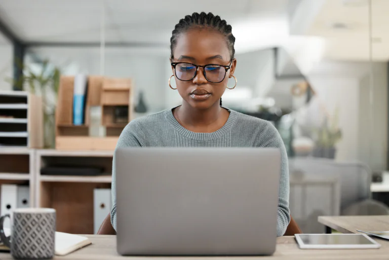 A woman wearing glasses and a grey jumper sitting at a desk working on a laptop.