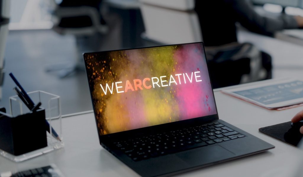 A laptop on a desk with an image displayed on screen. The image features rainbow smoke and says “WEARCREATIVE”.