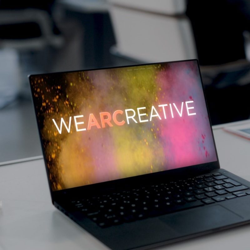 A laptop on a desk with an image displayed on screen. The image features rainbow smoke and says “WEARCREATIVE”.