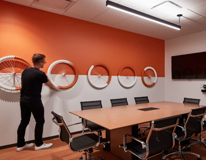 A man installing a wall feature on an orange and white horizontally split wall. The feature is 5 bike wheels painted orange and white.